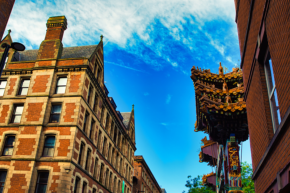 A low angle shot of a brick building with a ornate roof structure to the right. The sky is blue with white clouds in Manchester, UK.