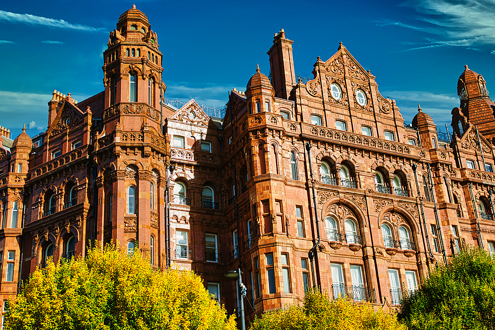 A red brick building with intricate details, multiple windows, and a clock face, viewed from a low angle. Trees frame the building, partially obscuring the view. The sky is blue with some clouds in Manchester, UK.