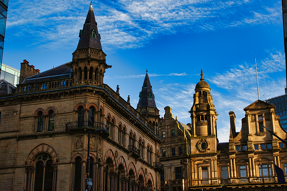 A low-angle shot of several ornate stone buildings with spires and intricate details against a bright blue sky with white clouds in Manchester, UK.