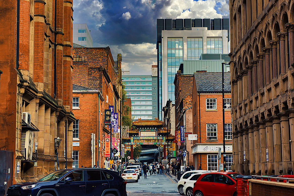 A narrow street lined with brick buildings, with a modern glass building at the end, and a Chinese archway in the middle. The sky is partly cloudy in Manchester, UK.