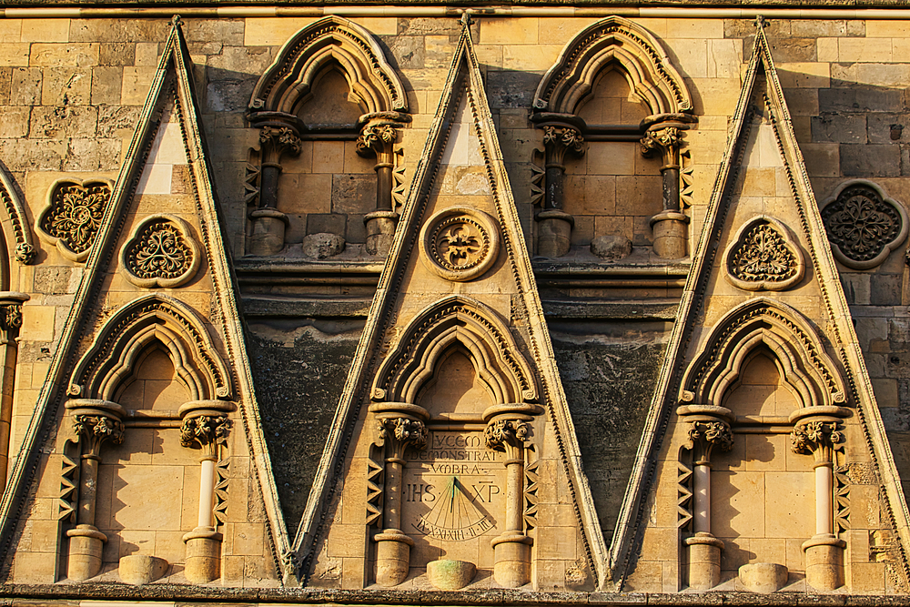 Close-up of a stone wall with intricate carvings. Triangular, arched windows with ornate columns and a sundial at the center in York, North Yorkshire, UK.