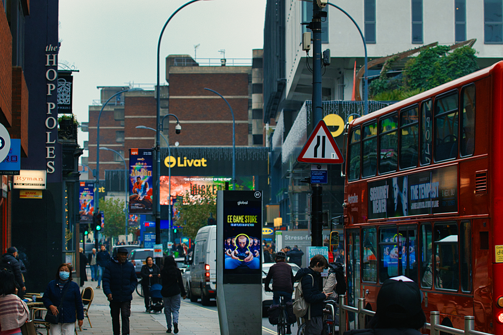 A busy city street with pedestrians, cars, and a double-decker bus. The bus is red and has advertisements on its side. People are walking along the sidewalk and crossing the street.