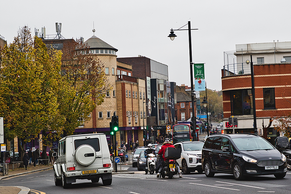 A street scene in an urban area, with a white SUV in the foreground and multiple vehicles, pedestrians, and buildings in the background.