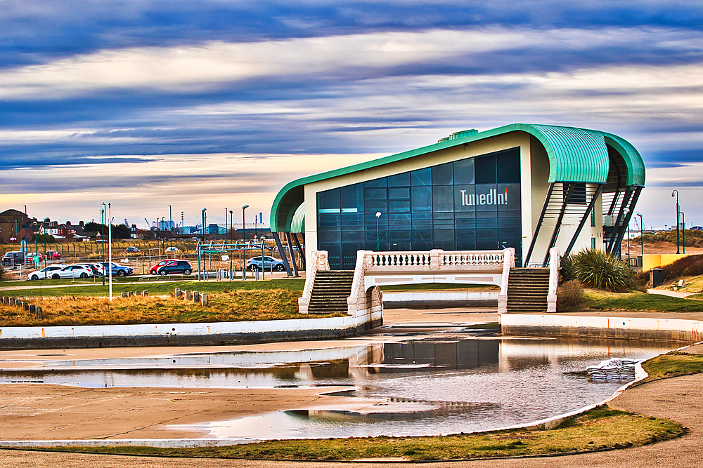 A modern, teal-roofed building with large glass windows and the word 'TunedIn!' visible, sits beside a small bridge over a calm, reflective water feature. Cars are parked in a lot in the background under a partly cloudy sky in Redcar, UK.