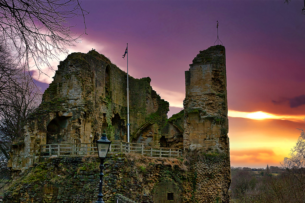 Ruins of a stone castle at sunset. The castle walls are partially collapsed, showing aged stonework. A flagpole stands within the ruins, and a lamppost is visible in the foreground. The sky displays vibrant orange and purple hues in Knaresborough, UK.