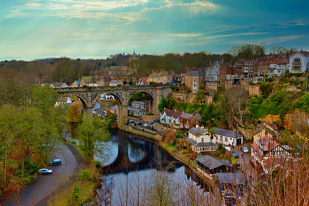 Stone viaduct over a river, with residential buildings and a church in the background. Trees line the river and the hillside. Cars are visible on a road below the viaduct in Knaresborough, UK.