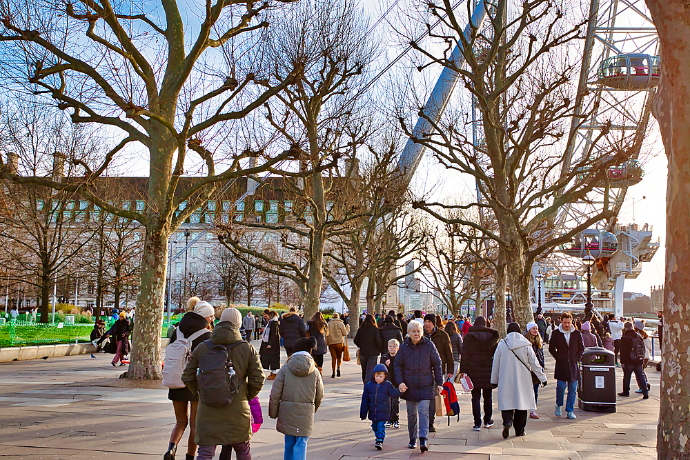 A crowd of pedestrians walks along a paved pathway lined with leafless trees. The London Eye is visible in the background. The scene is bright, suggesting daytime in winter.