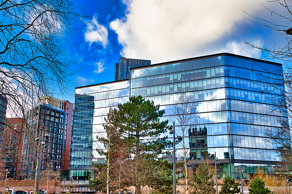 A modern glass-fronted building reflects a cloudy sky and surrounding structures. Trees and other buildings are visible in the reflection. The building is multi-storied with a dark frame in Manchester, UK.