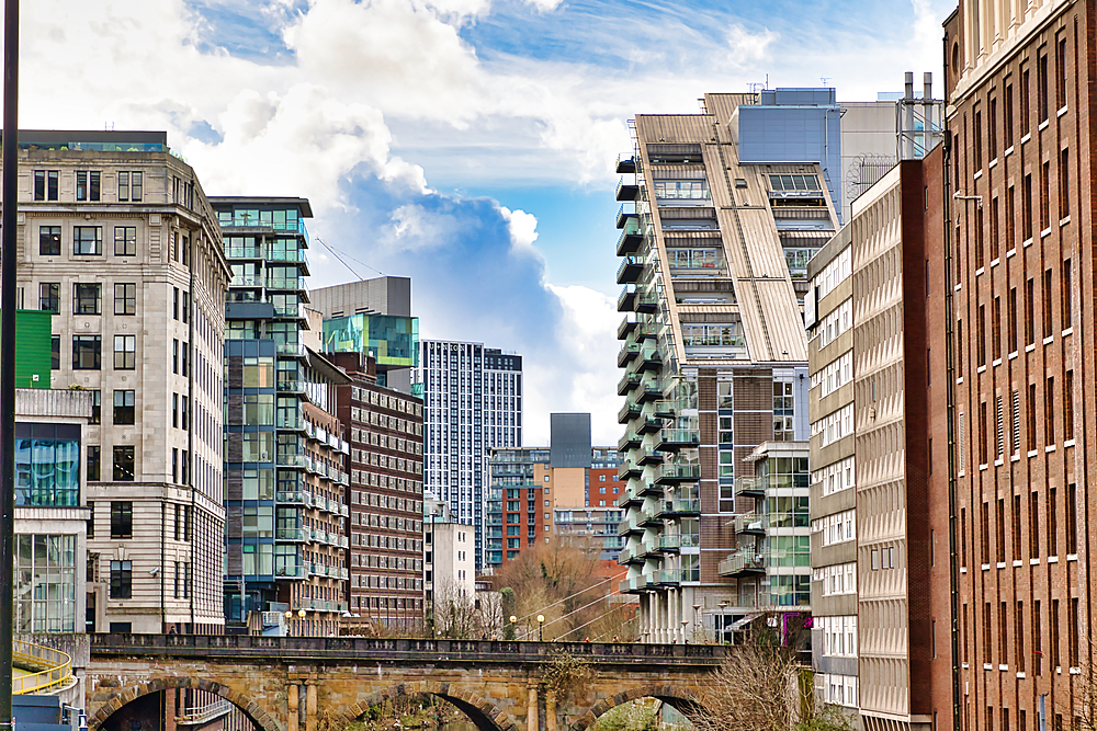 Modern and historic buildings line a waterway, viewed from a stone bridge. A slanted modern apartment building is a focal point, flanked by taller, older structures and a brick warehouse. Cloudy sky provides contrast in Manchester, UK.