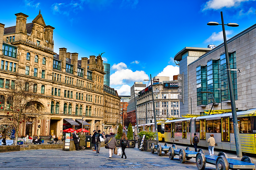 A sunny city street scene features a large, tan-colored Victorian-era building, modern glass structures, and a tram. Pedestrians are walking along the paved plaza. The sky is bright blue with some clouds in Manchester, UK.