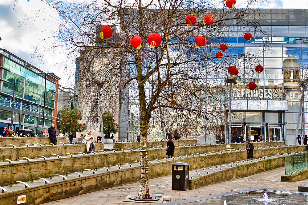 A city plaza features a tree adorned with red lanterns, stone seating, and a modern building reflecting the scene. People are visible, adding a sense of daily urban life in Manchester, UK.