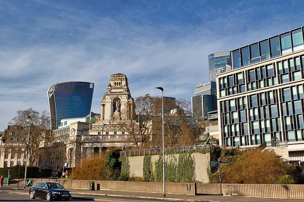 A cityscape featuring a mix of modern and classical architecture under a partly cloudy sky. A modern skyscraper stands beside a grand, classical building, with a contemporary building in the foreground. Cars and pedestrians are visible in London, UK.