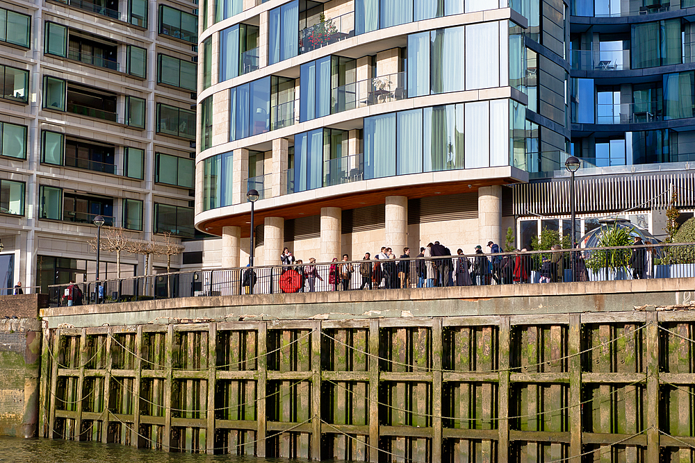 Modern apartment buildings and a group of people stand on a waterfront walkway above a weathered wooden retaining wall. The scene is brightly lit, showcasing architectural details and the people in London, UK.