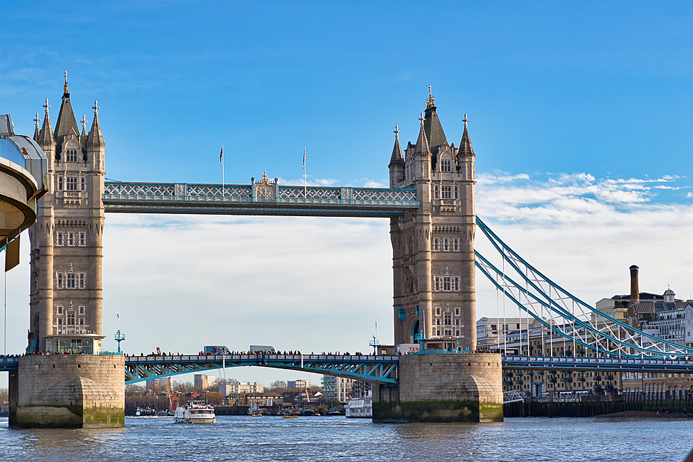 Tower Bridge in London, England, on a sunny day. The bridge is shown from a low angle, with people walking across. The River Thames and distant buildings are visible.