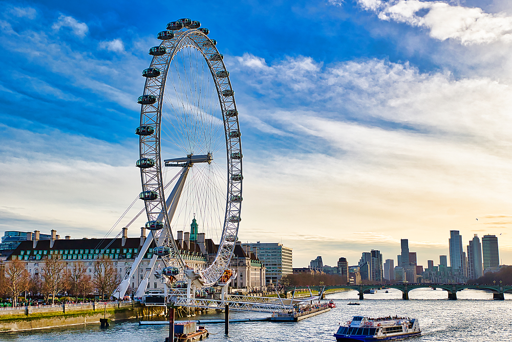 The London Eye Ferris wheel dominates this image, situated on the Thames River. Buildings line the riverbank, and a city skyline is visible in the distance under a partly cloudy sky. Several boats are on the water.