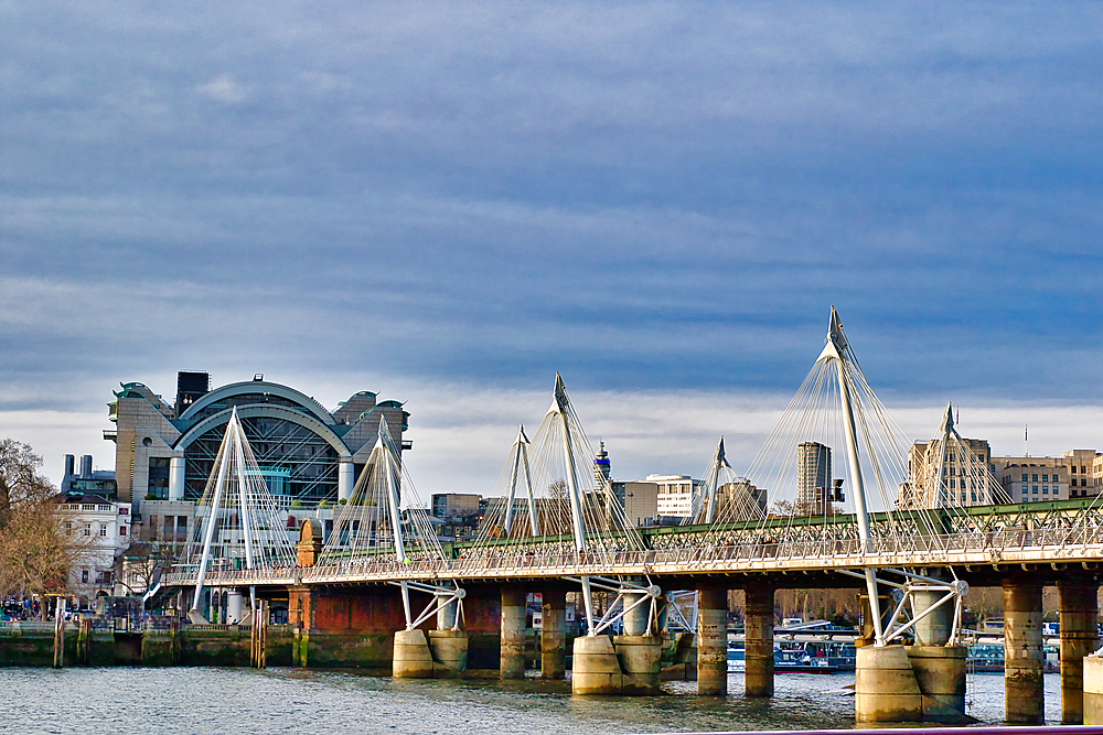 A cable-stayed pedestrian bridge spans a river, with a large modern building in the background under a partly cloudy sky. The scene is dominated by cool tones, with the bridge's white cables and grey structure contrasting against the water and sky in Lon