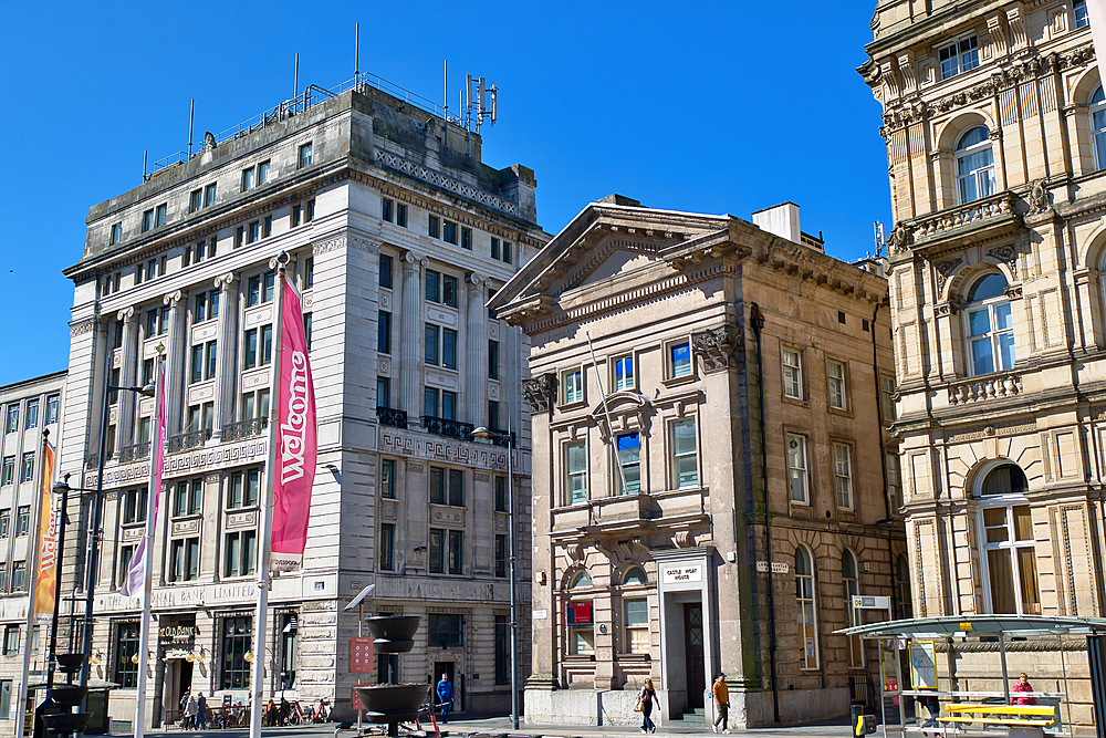Exterior shot of several buildings, including The National Bank, with people and street furniture, under a bright blue sky in Liverpool, UK.