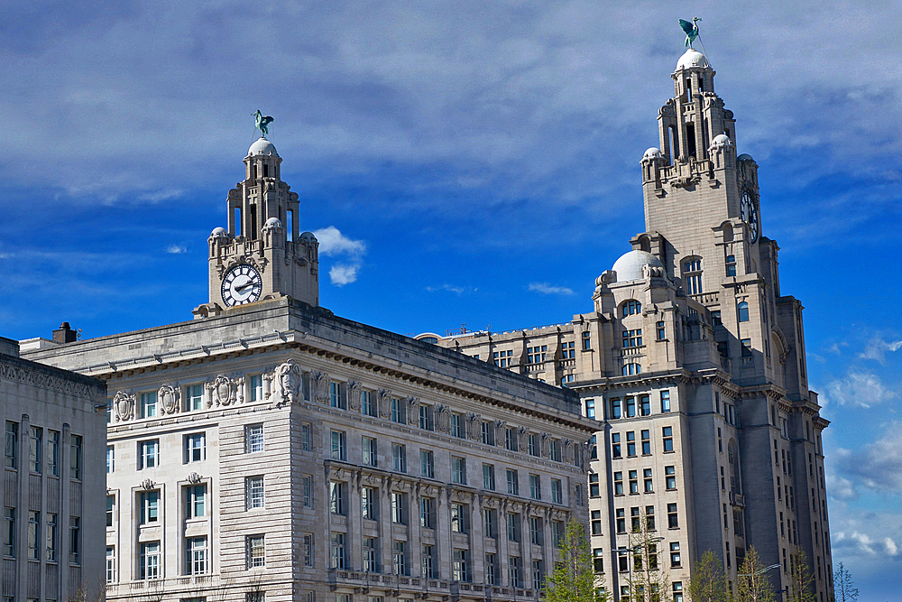 Architectural shot showcasing ornate, light stone buildings with clock towers topped by statues, set against a blue, cloudy sky in Liverpool, UK.