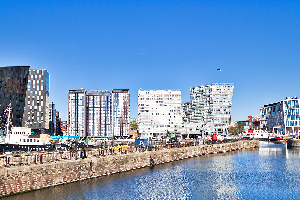 Urban waterfront view of modern buildings and docked ships under a clear blue sky. Features a brick dock wall in the foreground in Liverpool, UK.