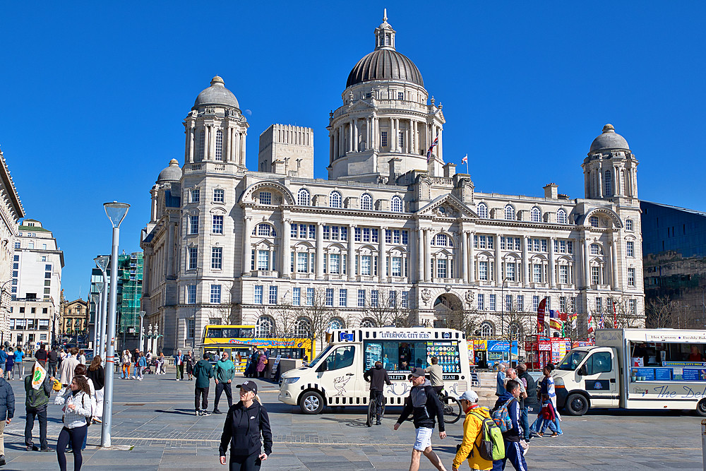 The image shows the Royal Liver Building in Liverpool, England, with people and food trucks in the foreground under a clear blue sky.