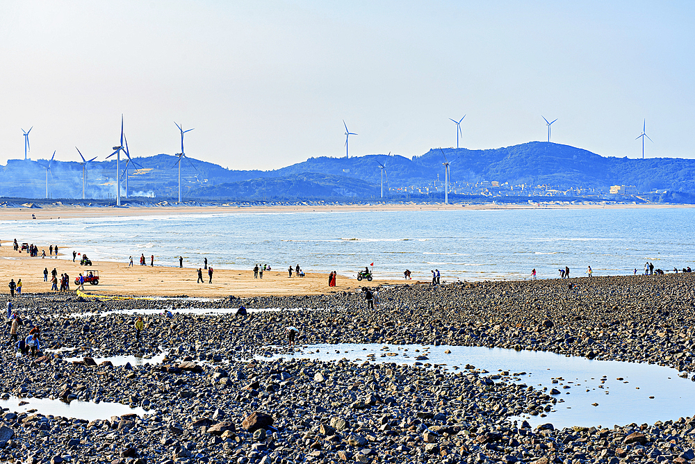 Contrast of rocky and sandy beach coastal recreation area with windmills in the background. Pingtan Island, Fuzhou City, Fujian Province, China.