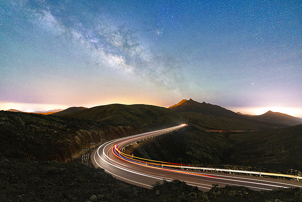 Milky Way glowing over the car trail lights on a winding desert road, Sicasumbre viewpoint, Fuerteventura, Canary Islands, Spain