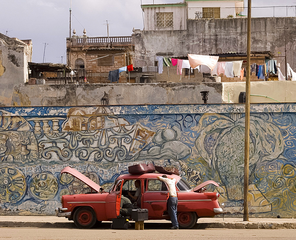 Two men repairing an old car parked in front of a colourful wall mural in central Havana, Cuba, West Indies, Central America