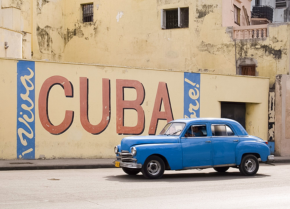 A vintage 1950's American car passing a 'Viva Cuba' sign painted on a wall in cental Havana, Cuba, West Indies, Central America