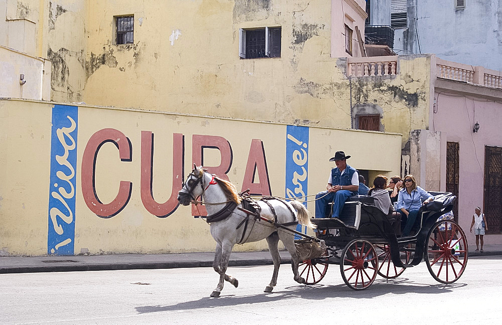 Tourists in a horse carriage passing a wall sign saying 'Live free Cuba', Havana, Cuba, West Indies, Central America