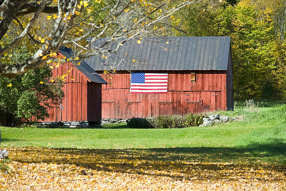 An old red barn with an American flag hanging on the side and autumn foliage on the hill behind, Vermont, New England, United States of America, North America