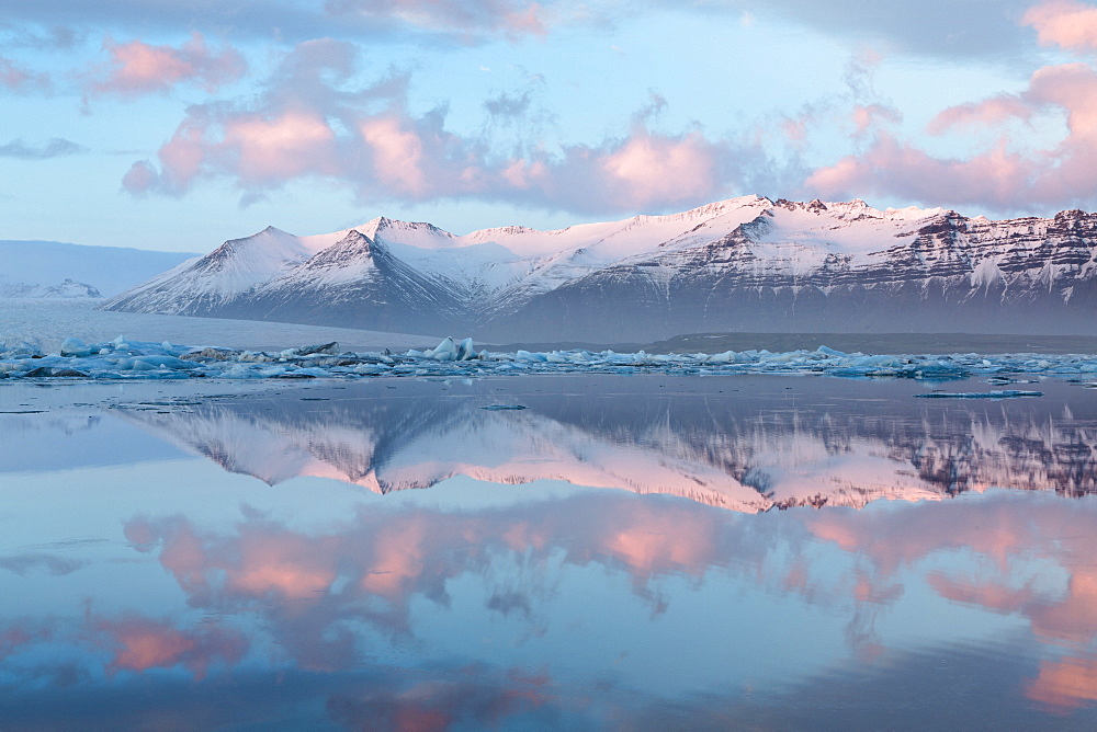 Panoramic view across the calm water of Jokulsarlon glacial lagoon towards snow-capped mountains and icebergs bathed in the last light of a winter's afternoon, at the head of the Breidamerkurjokull Glacier on the edge of the Vatnajokull National Park, South Iceland, Iceland, Polar Regions