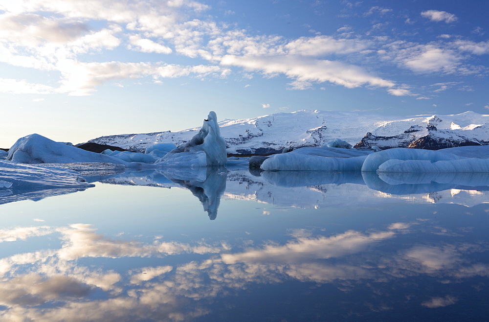 View over Jokulsarlon, a glacial lagoon at the head of the Breidamerkurjokull Glacier, towards icebergs and snow-capped mountains, with reflections in the calm water of the lagoon on a winter's afternoon, on the edge of the Vatnajokull National Park, South Iceland, Iceland, Polar Regions