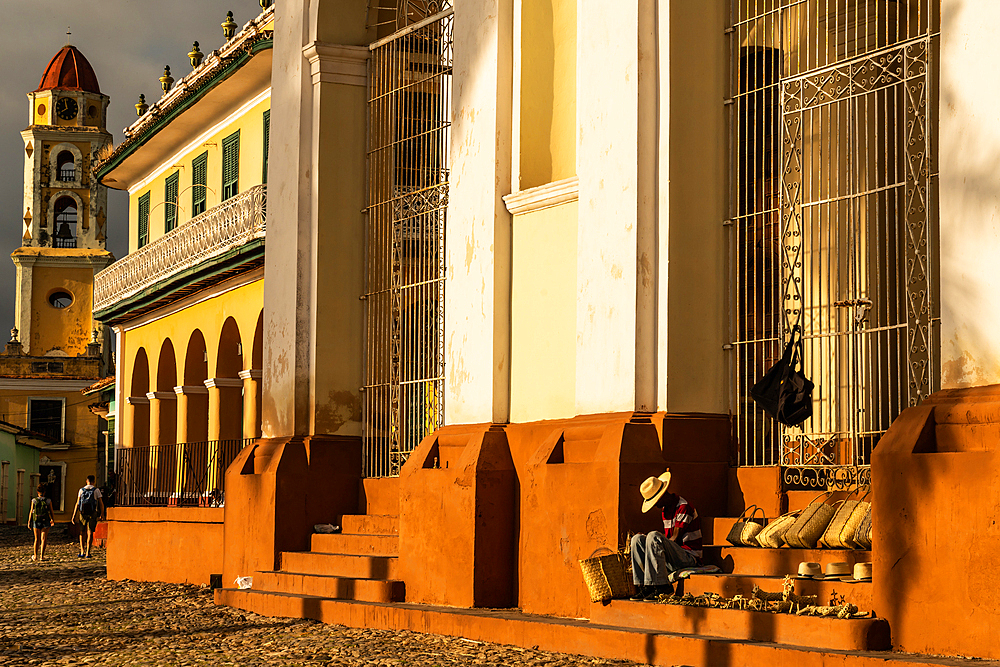 Evening sunlight bathes buildings of main square and souvenir seller on Cathedral steps, Trinidad, Cuba