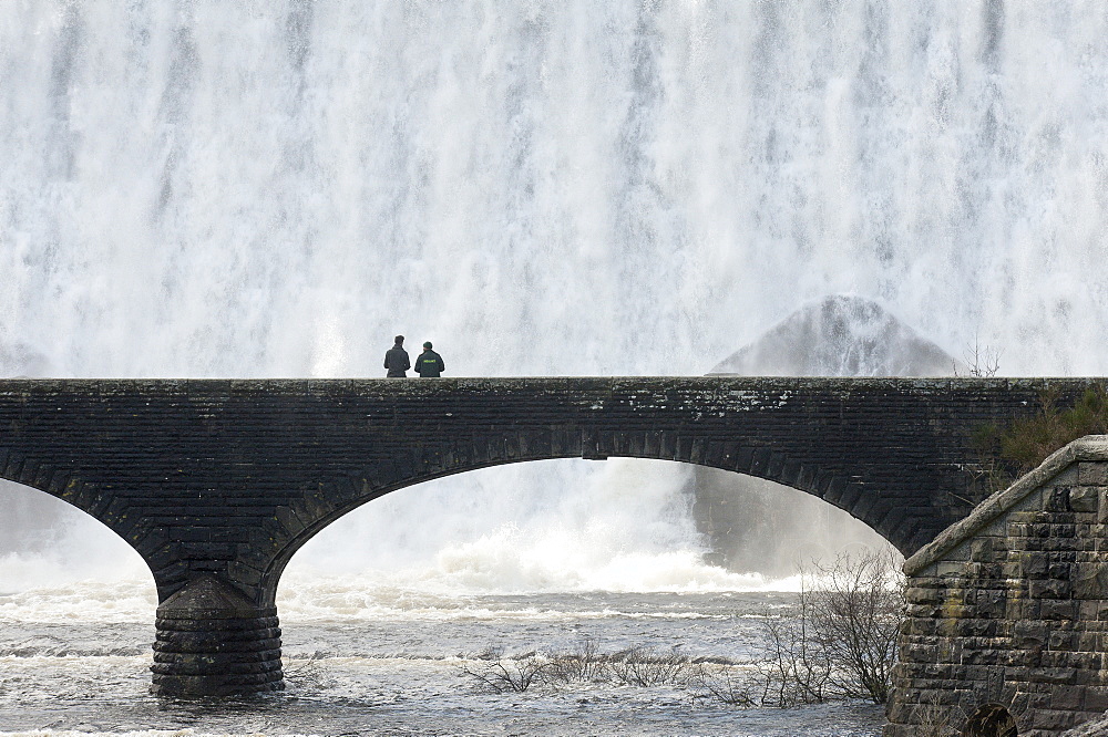 Visitors walk across a bridge as water cascades over the Caban-coch dam, at Elan Valley village near Rhayader in Powys, Wales, United Kingdom, Europe