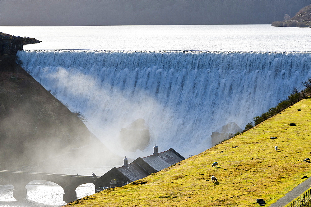 Visitors walk across a bridge as water cascades over the Caban-coch dam, at Elan Valley village near Rhayader in Powys, Wales, United Kingdom, Europe