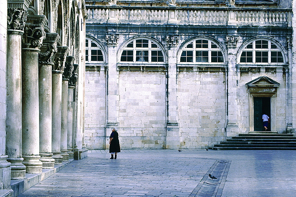 Croatia, Dalmatia, Dalmatian Coast, Fortified City Of Dubrovnik, Woman Walking On The Main Square, Duke's Palace On Left And Cathedral At Back