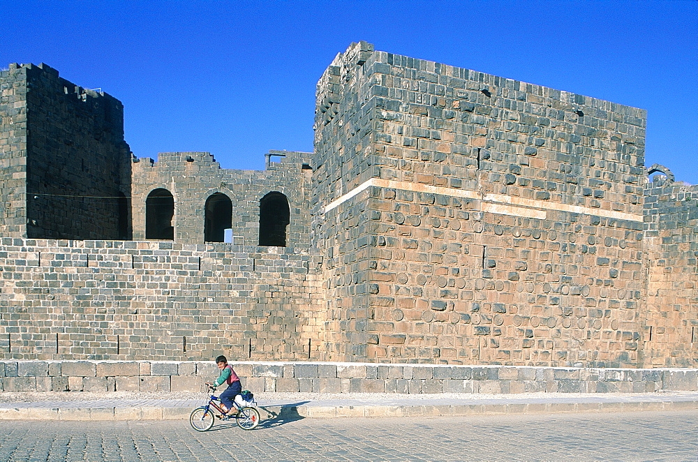 Syria, Haura, Bosra Ancient Roman Capital Of The Arabia Province, Child On A Bike Passing By The Fortess Walls, At Back The Theater