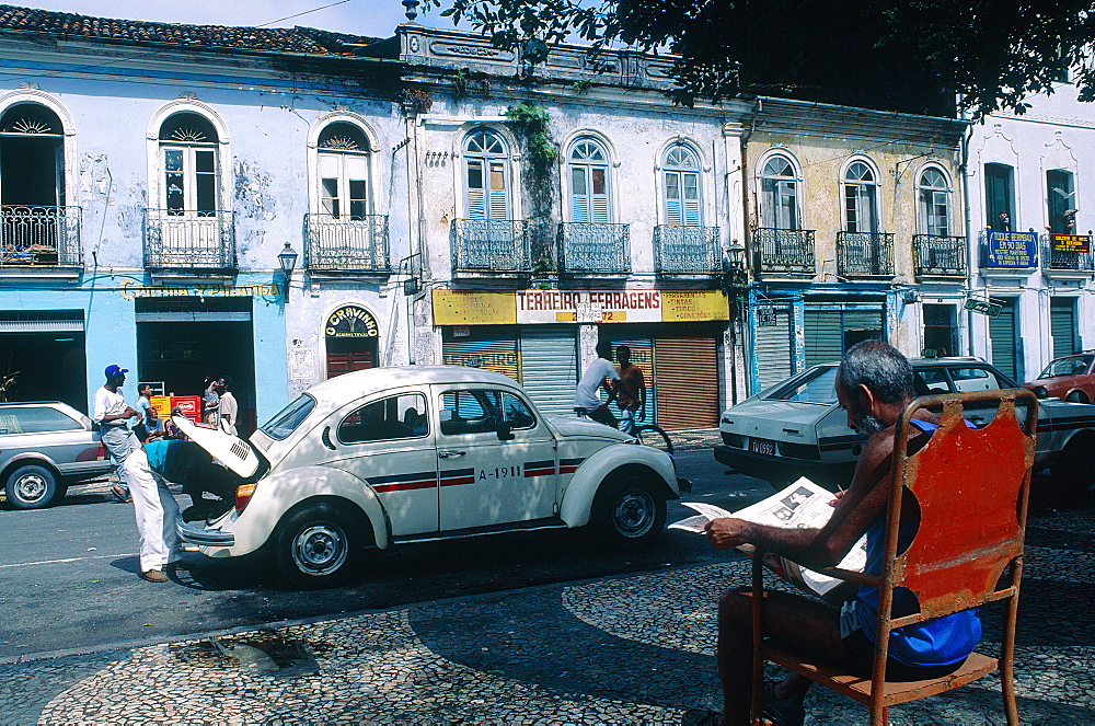 Brazil, Salvador De Bahia, The Historic Quarter Of Pelourinho, Man Fixing His Volskswagen Taxi Engine, On The Sidewalk A Man Sitting On A Big Armchair Reading A Paper