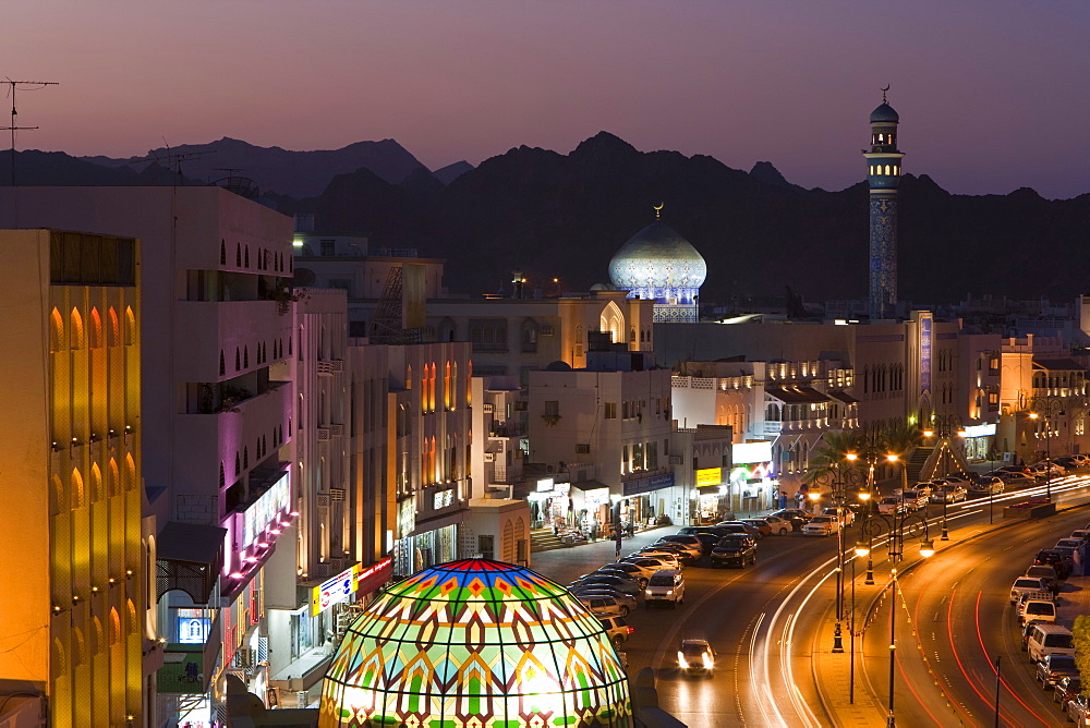 Elevated dusk view along the Corniche, stained glass dome of Mutrah Souq, colourful latticed buildings and Mutrah Mosque, Muscat, Oman, Middle East