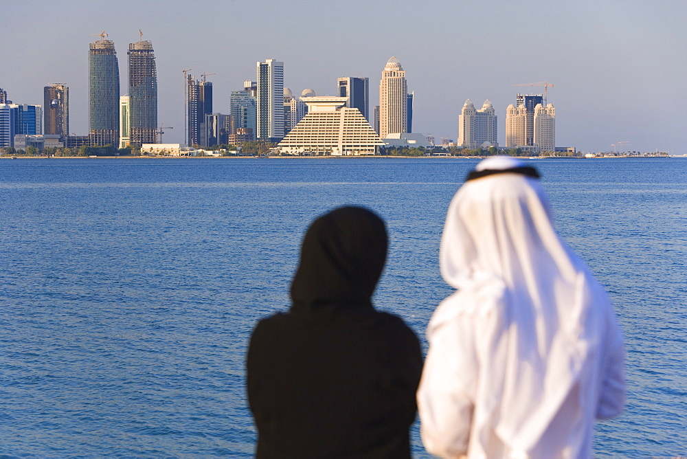 Man and woman in traditional dress looking across Doha Bay from the Corniche to the new city skyline and West Bay business and financial district, Doha, Qatar, Middle East