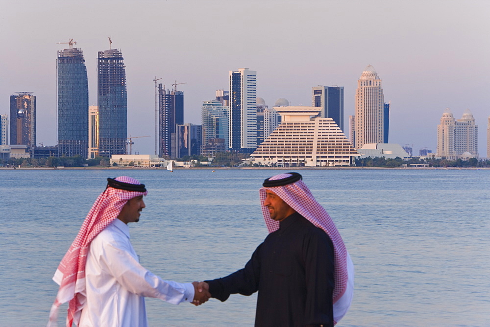 Two men wearing traditional dress of thobe, and gutra (headdress), shaking hands with Doha Bay, city skyline and West Bay business and financial district behind, Doha, Qatar, Middle East