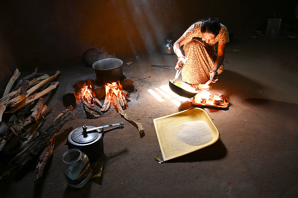 Sunlight streaming into traditional mud walled kitchen with wood fire, woman making chai, plastic rice sieve, Kadana, Gujarat, India, Asia