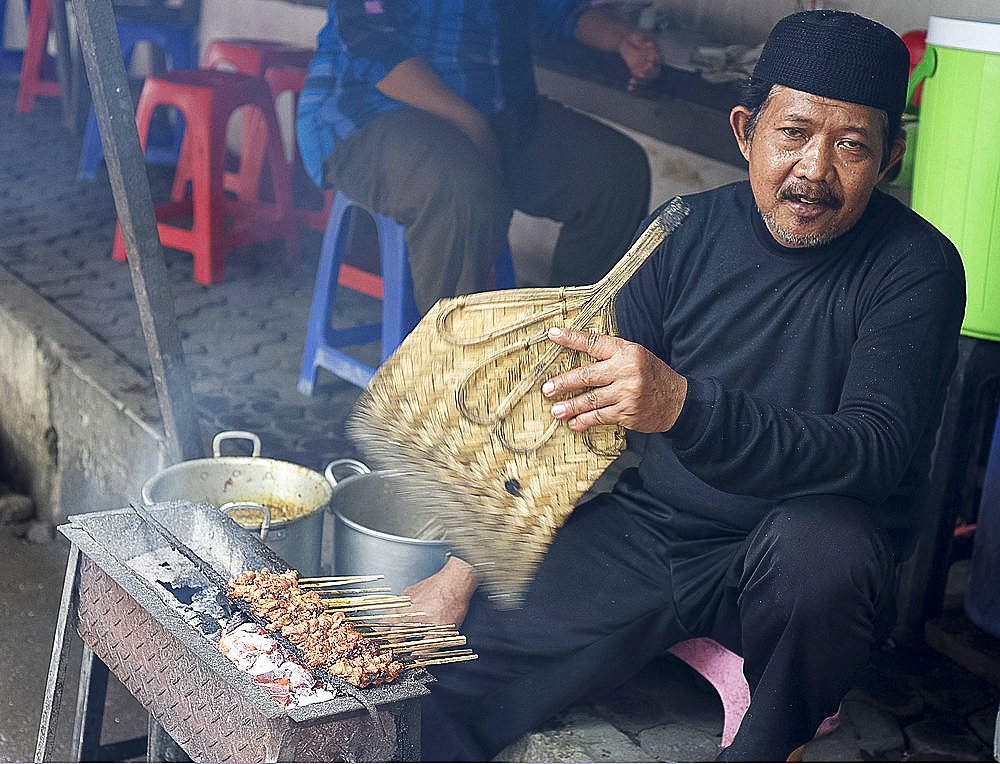 Muslim man in the market cooking chicken satay, fanning the charcoal with a palm leaf fan, Solo, Java, Indonesia, Southeast Asia, Asia