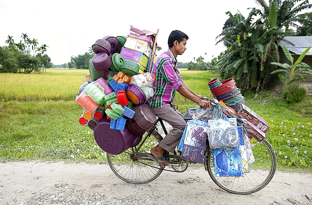 Young village tradesman cycling with a large load of plastic houseware, Majuli, Assam, India, Asia