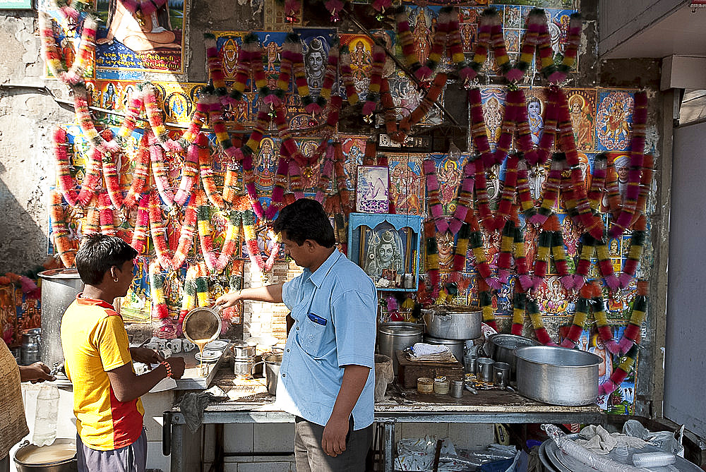 Chai wallah pouring chai (tea) at chai stall decorated with religious posters and malas (garlands), Ahmedabad, Gujarat, India, Asia