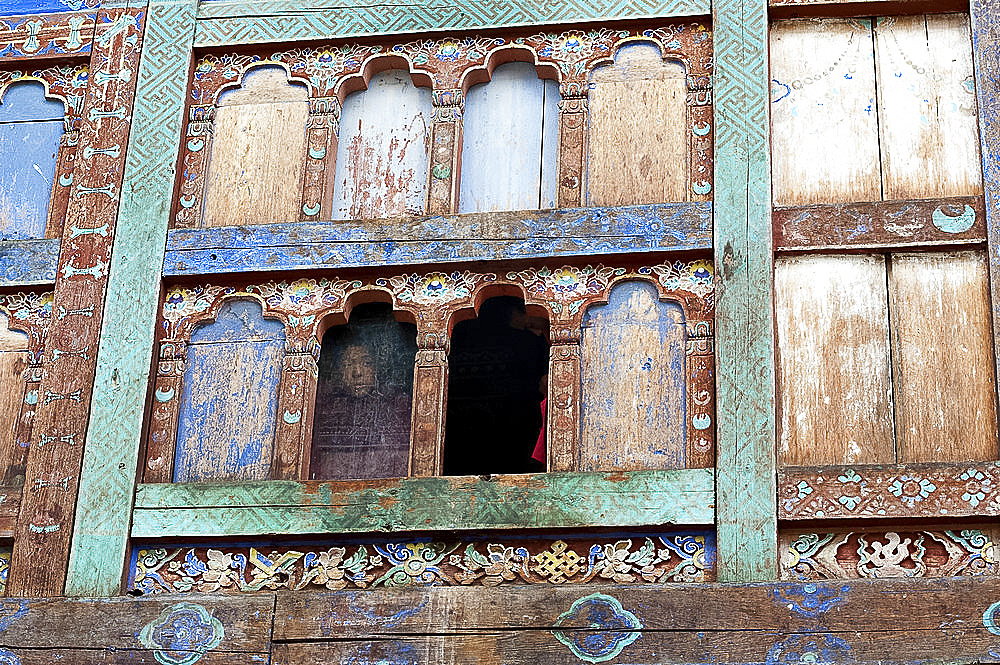 Monk looking out of window with fading blue paint in Wangdue Chhoeling Palace, in need of restoration, Bumthang, Bhutan, Asia