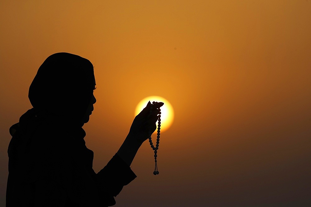 Silhouette of a Muslim woman holding prayer beads in her hands and praying at sunset, United Arab Emirates, Middle East