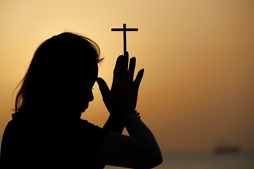 Silhouette of faithful woman praying with Christian cross at sunset as concept for religion, faith, prayer and spirituality, Dubai, United Arab Emirates, Middle East