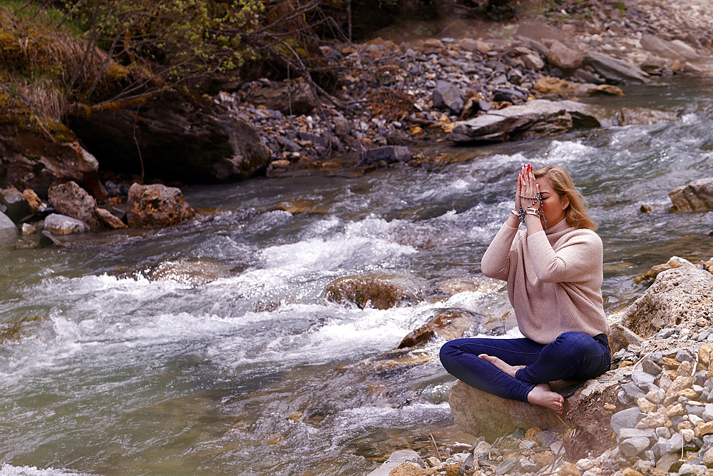 Praying woman sitting on a rock at the edge of a river, concept of religion in nature, Les Contamines, Haute-Savoie, France, Europe