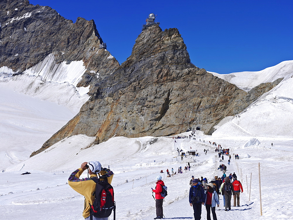 People walking on the Great Aletsch Glacier, the longest glacier in the Alps, begins on the Jungfraujoch (Top of Europ), with The Sphinx building on the top of the mountain, Switzerland, Europe